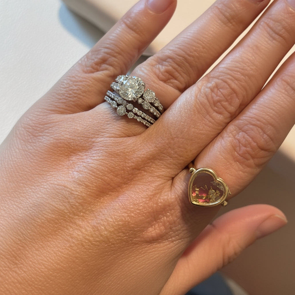 Hand wearing two diamond rings with a heart-shaped gold ring featuring a colorful gemstone.